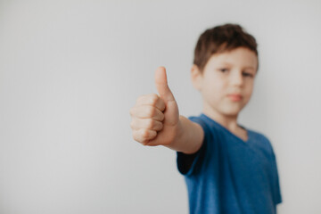 A preschool boy in a blue T-shirt on a light background shows a thumbs up