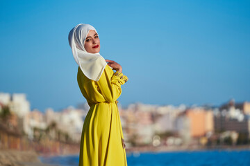 Arab woman, on a beach walking along the shore posing, wearing typical Moroccan yellow clothing and a white hijab covering her face smiling. Horizontal photo