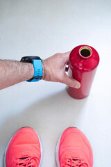 a hand with a blue sports smartwatch picks up a red water bottle from the floor during a break in a workout session at the gym, pink sneakers are seen in the foreground, vertical