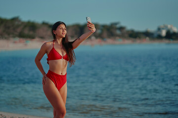 Young Latina girl on the beach enjoying the summer in a red bikini, taking a selfie with her cell phone. Horizontal photo
