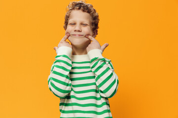 horizontal portrait of a cute, funny, playful, curly-haired boy of school age in a striped long-sleeved t-shirt on an orange background pulling a smile with his fingers