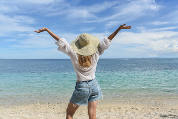 A woman is resting on the beach. She is wearing a straw hat, denim shorts and a white shirt. Travel and good rest, happy holidays, tourism, Philippines, summer season. Rear view.