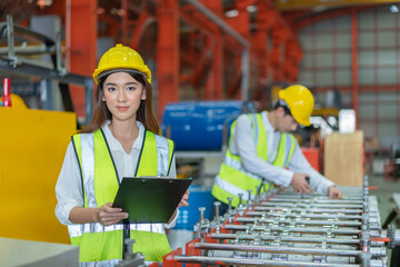 Young female engineer wearing safety vest with yellow helmet checking machine on document in factory
