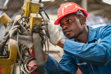 African American male engineer in red safety vest and helmet checking and repairing old robot arm in factory Industrial