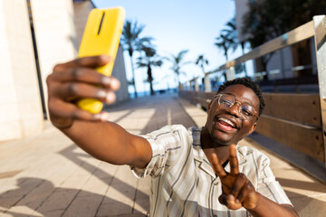 Young black man taking selfie using phone making peace sign. Happy, African american male having fun. - obrazy, fototapety, plakaty