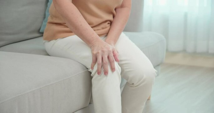 Close up of elderly Asian woman suffering from pain in knee while getting up from the sofa in the living room at home.Old female with osteoarthritis.Health problem and people concept.