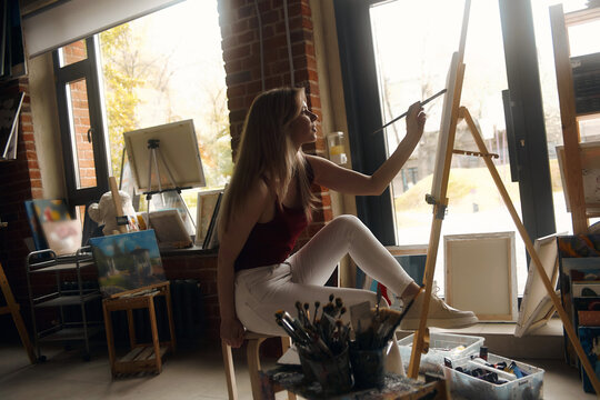 Inspired blonde woman with a brush, in light clothes smiling in the interior of the studio. Creative female student enjoying her new hobby, painting still lifes, side view. Art concept