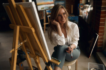 Inspired blond teenage girl with brush and palette smiling in studio interior. Creative schoolgirl enjoying her new hobby, still life painting, side view. Art concept