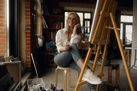 Inspired blond teenage girl with brush and palette smiling in studio interior. Creative schoolgirl enjoying her new hobby, still life painting, side view. Art concept