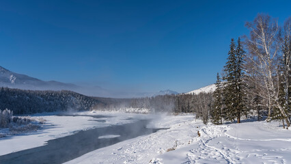 Fototapeta na wymiar An ice-free river in a snow-covered valley. Steam rises above the water. Footprints are visible in snowdrifts, trees on the shore. Mountains against the blue sky. Altai