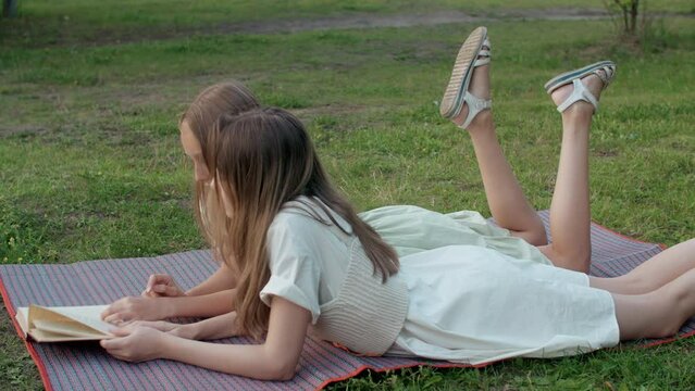 Two cute teenage girls spend weekend or vacation reading book lying on green grass and swing legs in park outdoors on summer day. Side view