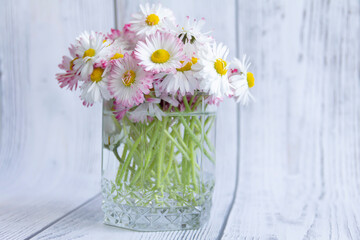 A bouquet of fresh flowers, daisies on green stems stand in a glass with clean water on a gray, wooden background.