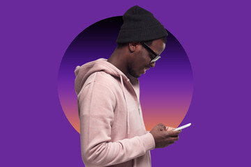 Side portrait of smiling black guy in hat and shades using mobile phone over purple background