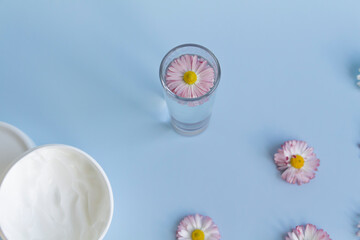 Fresh flowers, daisies are laid out in a corner, with a glass of clean water and a white jar with a cream cosmetic on a blue background and a free space for advertising.