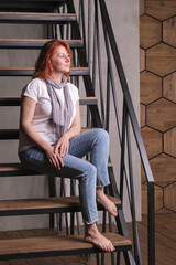 Red-haired attractive woman sits on a metal staircase. A woman in jeans and barefoot looks away and thinks about her future. Vertical portrait of a woman in full length profile.