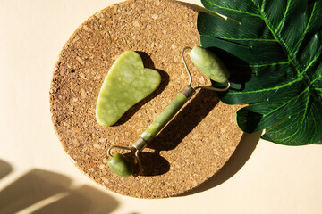 Jade Gua sha scraper and face roller massager on a cork round stand with a monstera leaf.