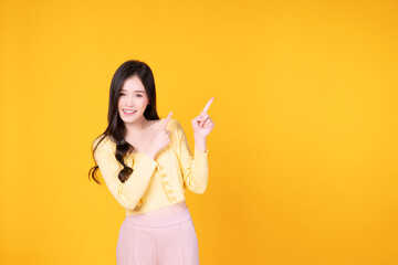 Cheerful Asian woman use finger pointing to product or empty copy space standing over isolated on yellow background. Surprised model young girl present content. Advertisement presenting concept.