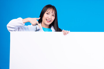 Positive cheerful asian woman showing white placard empty copy space standing over isolated blue background. Teenager girl pointing to blank Banner, advertisement, sign board presentation concept.