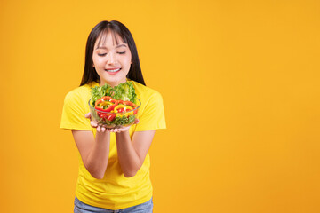 Cheerful Asian woman slim body holding glass bowl with organic fresh vegetables for good health diet standing over isolated yellow background. Fit body young girl Health care lose weight food concept.