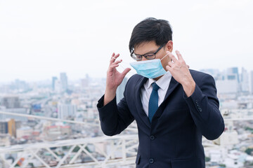 Asian businessman with eyeglasses wearing face mask protection air pollution or covid-19 on cityscape background. Business young man wear suit with surgical mask standing alone on root top building.