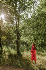 Pretty young redhead woman in red dress walking through meadow with flower in hand. Low angle view portrait of smiling female person on the forest bckground