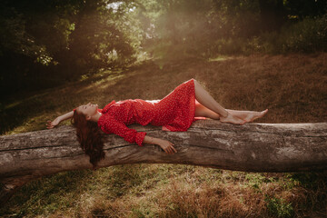 a young and beautiful woman in a red summer dress with white flecks lying on a fallen dry tree...