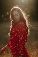 Medium shot portrait of redhead young woman in red dress with white dots in sunset backlight background. Female model looking at camera