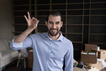 Happy millennial guy showing bunch of keys on camera, standing in living room with belongings in...