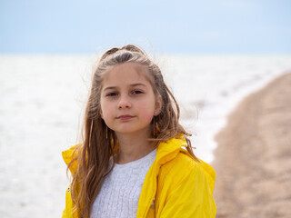 A pretty girl with long hair stands against the background of the water surface.  Open portrait of a smiling blonde teenage girl in a yellow windbreaker on the seashore