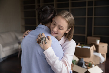 Millennial couple hugging standing in living room with boxes on relocation day. Attractive young wife holding bunch of keys embracing her husband enjoy move-in. Bank mortgage, tenancy, love concept