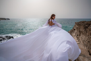 Happy freedom woman on the beach enjoying and posing in white dress. Rear view of a girl in a fluttering white dress in the wind. Holidays, holidays at sea.