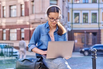 Teenage student girl in headphones using laptop outdoor, urban background