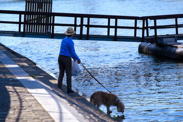 Man with stay hat and blue shirt let his dog drinking water from Rhine River on a sunny spring morning at City of Basel. Photo taken May 11th, 2022, Basel, Switzerland.