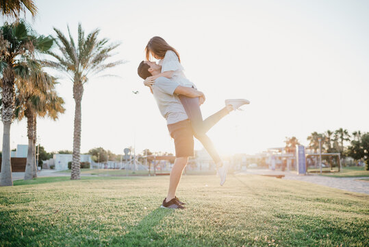 A full-length photo of a Hispanic man who is lifting his happy brunette girlfriend between palm trees and sunlight in Spain in the evening. A couple of tourists on a date at the sunset in Valencia.