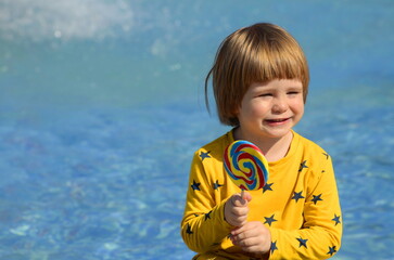 A little boy is enjoying a big candy on a stick. Multicolored lolipop. Child on a blue background....