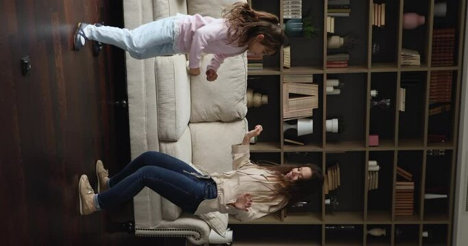Vertical view young lively mother her little cute daughter holding hands dance, moving to favourite music having fun fooling at modern fashionable living room. Funny activity with kids at home concept
