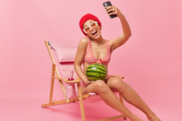 Cheerful happy woman has good mood makes selfie via smartphone while poses on deck chair holds...
