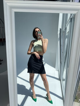 Fit tanned woman in classic short black skirt, light green crop top and heels, summer fashion stylish wear, take photo selfie on phone in mirror for social media, stories, vertical.