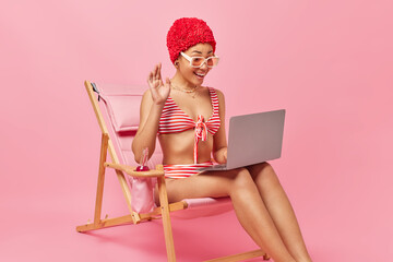 Positive woman waves hand and talks on video chat via laptop computer during livestream uses high speed internet for good connection dressed in swimwear sits on deck chait enjoys summer vacations