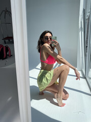 Fit tanned woman in light green beach skirt, sunglasses, bright pink swimsuit bra, summer fashion stylish wear, take photo selfie on phone in mirror for social media, stories, vertical.