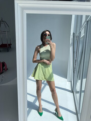 Fit tanned woman in light green beach skirt, sunglasses, crop top and heels, summer fashion stylish wear, take photo selfie on phone in mirror for social media, stories, vertical.