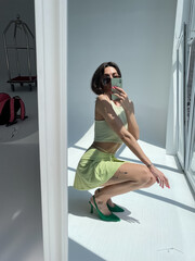 Fit tanned woman in light green beach skirt, sunglasses, crop top and heels, summer fashion stylish wear, take photo selfie on phone in mirror for social media, stories, vertical.