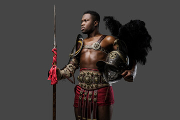 Portrait of muscular african gladiator holding feathered helmet and spear isolated on gray background.