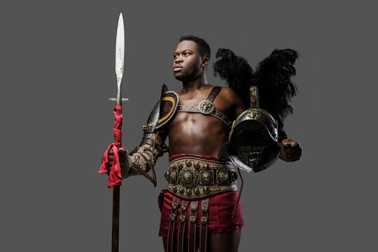 Portrait of muscular african gladiator holding feathered helmet and spear isolated on gray background.