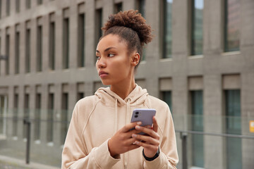 Fototapeta na wymiar Pensive female trainer dressed in hoodie uses smartphone for messaging online gives sportive advice has break on workout downloads music for active training poses against city building background