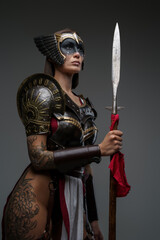 Shot of antique female warrior dressed in steel armor holding spear against grey background.