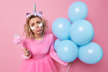 Fototapeta na wymiar Unhappy frustrated woman cries and has bad mood during party holds magic wand bunch of inflated blue helium balloons wears festive dress applies beauty patches isolated over pink background.