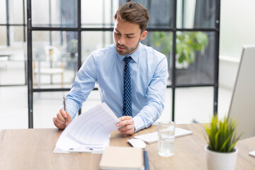 Young man is working with papers while sitting in the office. Successful entrepreneur is studying documents with attentive and concentrated look.