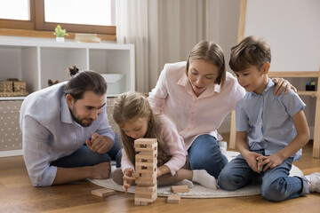 Interested children and parents engaged in wooden board game sit on floor in nursery, have fun entertaining at home. Playtime, carefree leisure, weekend together. Offspring growth, development concept