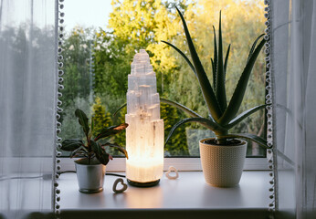 Rough big selenite crystal tower pole lamp illuminated in home on window sill, summer forest on...